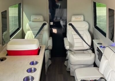 Interior of indulger specialty vehicle showing 2 white bucket seats, cooler, bench seats, and kitchenette on left side.