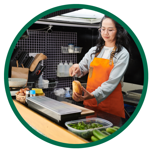 Round image with green outside circle of woman in orange apron working in food services trailer cutting bread.