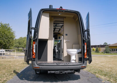 Back end of black TWR specialty vehicle with doors open, exposing a toilet seat and cabinet on the inside.