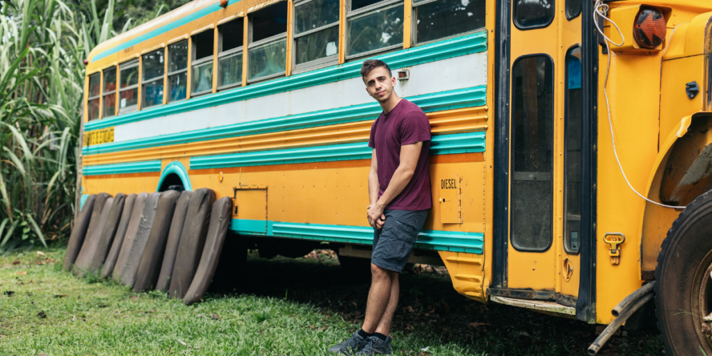 A young man leaning against a school bus in the midst of a skoolie conversion.